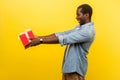 Take this present! Side view of generous man in denim casual shirt holding out red gift box. indoor studio shot isolated on yellow Royalty Free Stock Photo
