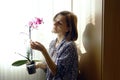 A young woman holds a purple orchid in her hands, standing in a room against the background of a window