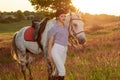 Jockey young girl petting and hugging white horse in evening sunset. Sun flare