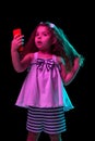 Taking selfie. Stylish little girl, beginner fashion model using smartphone isolated over dark background in neon light Royalty Free Stock Photo