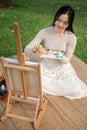 A talented Asian female artist is mixing water color on a palette, painting on a canvas in a park