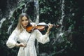 Talented Caucasian woman playing violin near waterfall. Music and art concept. Charming female with long blond hair wearing white Royalty Free Stock Photo