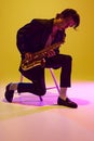 Talented jazz man, virtuoso playing saxophone sitting on chair in neon light against vibrant yellow studio background.