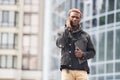 Talking by phone. Young african american man in black jacket outdoors in the city standing against modern business building Royalty Free Stock Photo