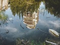 A tall abandoned building surrounded by a forest is reflected in the green water of a pond overgrown with grass and