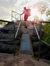 Tall backpacker hold handrail on rock. Sunny daybreak in rocks. Hiker with big backpack, baseball cap, dark pants and white shirt Royalty Free Stock Photo