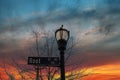 A tall black metal lamp post with street signs surrounded by bare winter trees with blue sky and powerful red clouds