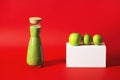 A tall bottle of green smoothies stands on a red background, next to it lies a white geometric prism with a green apple