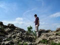 A tall dark-haired young man stands on a rock and looks down - view from the back. Clear sunny day, stone boulders and one adult