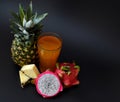 A tall faceted glass of fresh fruit juice on a black background, next to pieces of ripe pineapple and half a pitaya Royalty Free Stock Photo