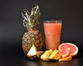 A tall faceted glass of fruit juice on a black background, next to a ripe pineapple, half a grapefruit and mango pieces Royalty Free Stock Photo