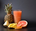 A tall glass of mixed fruit juices on a black background, next to pieces of ripe pineapple, mango and grapefruit Royalty Free Stock Photo