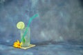 A tall glass of mojito with ice stands on a textile stand next to a vanilla flower bud the whole composition on a gray abstract