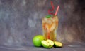A tall glass of pale yellow fruit juice with ice and straws stands against a gray background next to several green apples