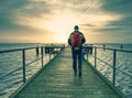 Tall man in the warm jacket and black pants is walking on the waterfront and looking at the sea. The wood pier on shore of sea