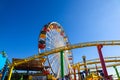 A tall metal Ferris wheel with colorful cars around the wheel on with a yellow rollercoaster rail and blue sky