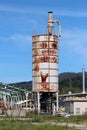 Tall partially rusted metal storage silo surrounded with destroyed buildings at abandoned industrial complex surround with uncut