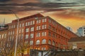 A tall red brick building with some boarded up windows on the street with a blue sky and powerful red and yellow clouds at sunset Royalty Free Stock Photo