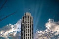 A tall slender skyscraper surrounded by bare winter trees in downtown with a blue sky and powerful clouds with sunrays