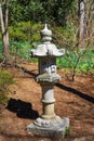 A tall slender stone Japanese lantern in the garden surrounded by brown fallen pine needles and lush green trees and plants Royalty Free Stock Photo