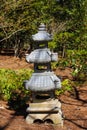 A tall slender stone Japanese lantern in the garden surrounded by brown fallen pine needles and lush green trees Royalty Free Stock Photo