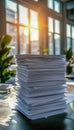 A tall stack of paper documents on a desk in a bright office with sunlight streaming through large windows Royalty Free Stock Photo
