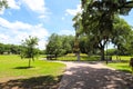 A tall tower shaped monument in the park surrounded by lush green grass and weeping willow trees with blue sky and clouds