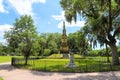 A tall tower shaped monument in the park surrounded by lush green grass and weeping willow trees with blue sky and clouds