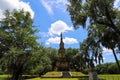 A tall tower shaped monument in the park surrounded by lush green grass and weeping willow trees with blue sky and clouds