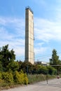 Tall and wide concrete industrial chimney with protective metal fence on top surrounded with abandoned factory buildings and overg