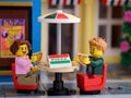 Two Lego minifigures sitting behind a table and eating pizza outdoors