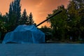 a tank covered with material stands in a city park against the background of an evening orange sky