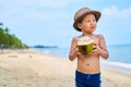 Tanned Asian boy stands on the beach in a hat and drinks coconut