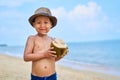 Tanned Asian boy stands on the beach in a hat and drinks coconut