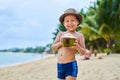 Tanned Asian boy stands on the beach in a hat and drinks coconut