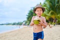 Tanned Asian boy stands on the beach in a hat and drinks coconut