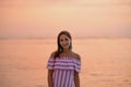 Tanned beautiful woman in dress with white and pink stripes. Orange sea or ocean at the sunset. Vacations concept Royalty Free Stock Photo