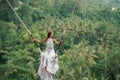 Tanned beautiful woman in a long white dress with a train, riding on a swing. In the background, a rainforest and palm trees. Copy