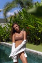 Tanned brunette in beige bikini and white shirt standing  under sun near pool surrounded with tropical plants at villa Royalty Free Stock Photo