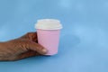 Tanned hand of elderly woman holds pink paper coffee cup closed with white lid on blue background. Natural eco packaging