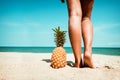 Tanned legs of young woman standing with pineapple at tropical beach in summer