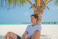 Tanned man wearing white t-shirt and black shorts relaxing under palm tree at tropical sandy beach at island luxury resort Royalty Free Stock Photo