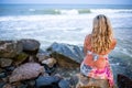 A woman with blond hair flying in the wind sits on boulders near the sea in a bright summer shawl Royalty Free Stock Photo