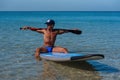 Tanned sporty man in a cap sits on his surfboard on the water holding by hands an oar behind his head and looks into the