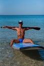 Tanned sporty man in a cap sits on his surfboard on the water holding by hands an oar behind his head and looks into the