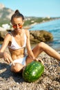 Tanned woman in white bikini and red glasses cuts with a knife huge ripe watermelon Royalty Free Stock Photo