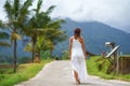 A tanned woman in a white dress walks forward on the road. The view from the back. In the background, a mountain in the fog and Royalty Free Stock Photo