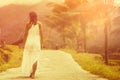 A tanned woman in a white dress walks forward on the road. The view from the back. In the background, a mountain and palm trees. Royalty Free Stock Photo