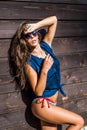 Tanned young woman with sunglasses in bikini and blue summer shirt by wooden house summer day