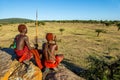 Two young Masai warriors are sitting on a big stone in traditional clothes with a spear in the savannah against a blue sky.